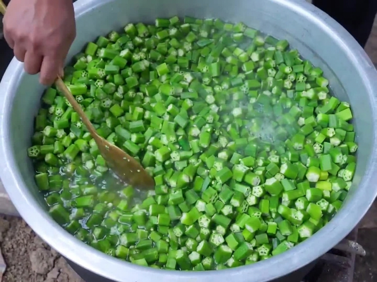 Chopped green okra being blanched in boiling water inside a large pot while being stirred with a wooden spoon.