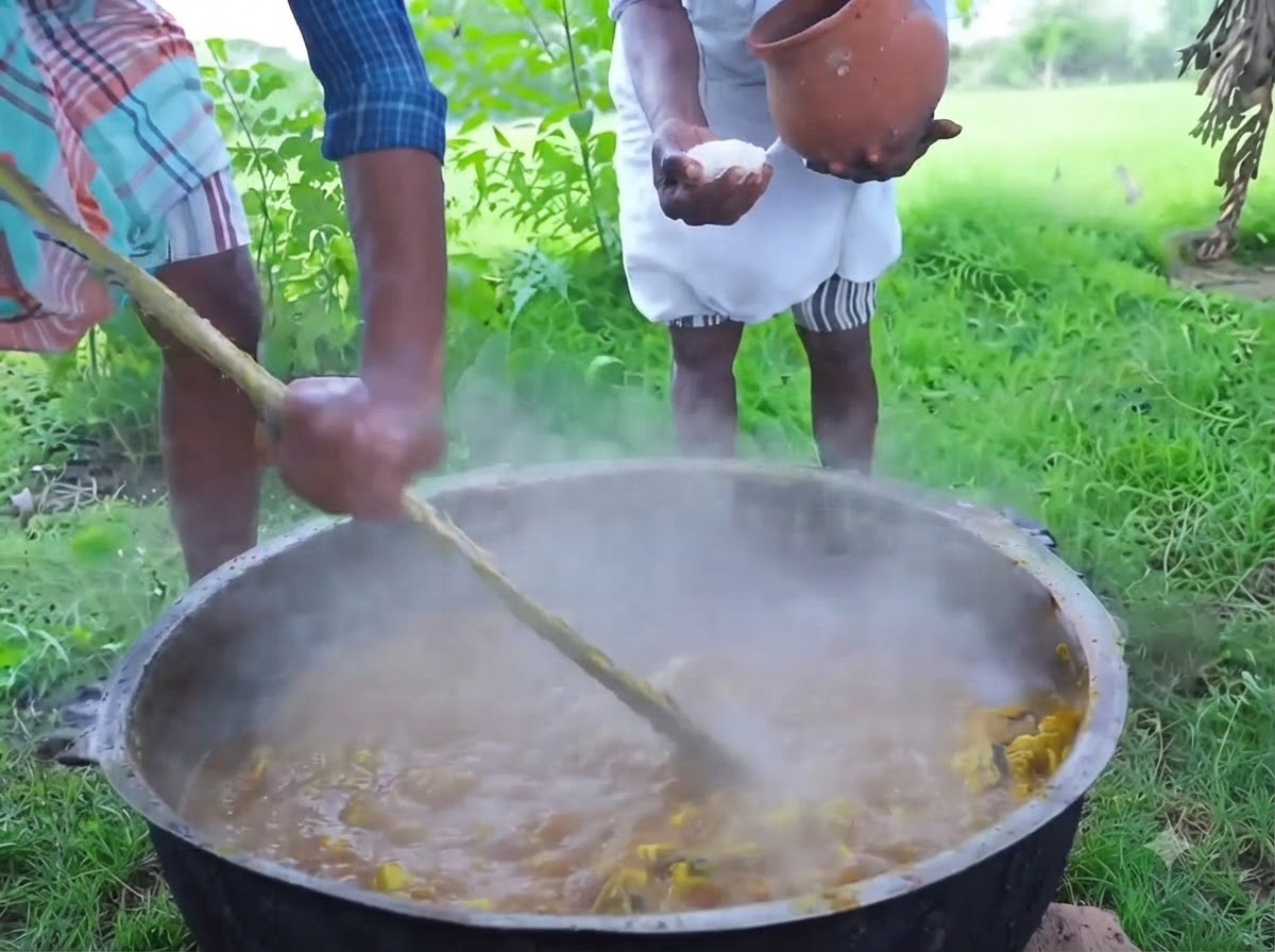 A person pouring white crystal salt from a clay pot over a steaming mixture in a large iron pot.