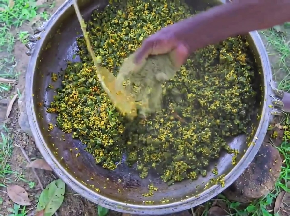 A persons hand mixing black pepper powder into a large iron pot filled with cooking okra and eggs.
