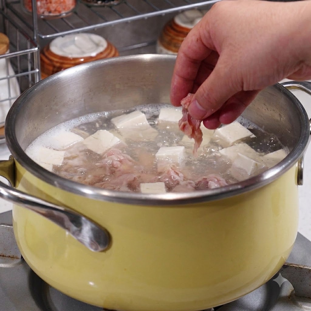 A hand placing marinated pork slices into a pot containing boiling broth, tofu, and seafood.
