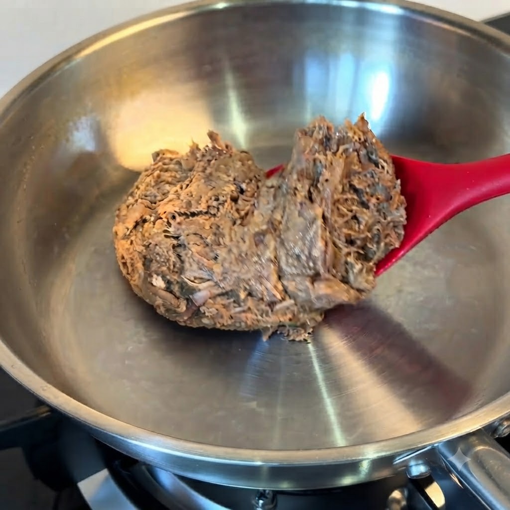 A block of thawed pulled beef being placed into a stainless steel pan using a red spatula.