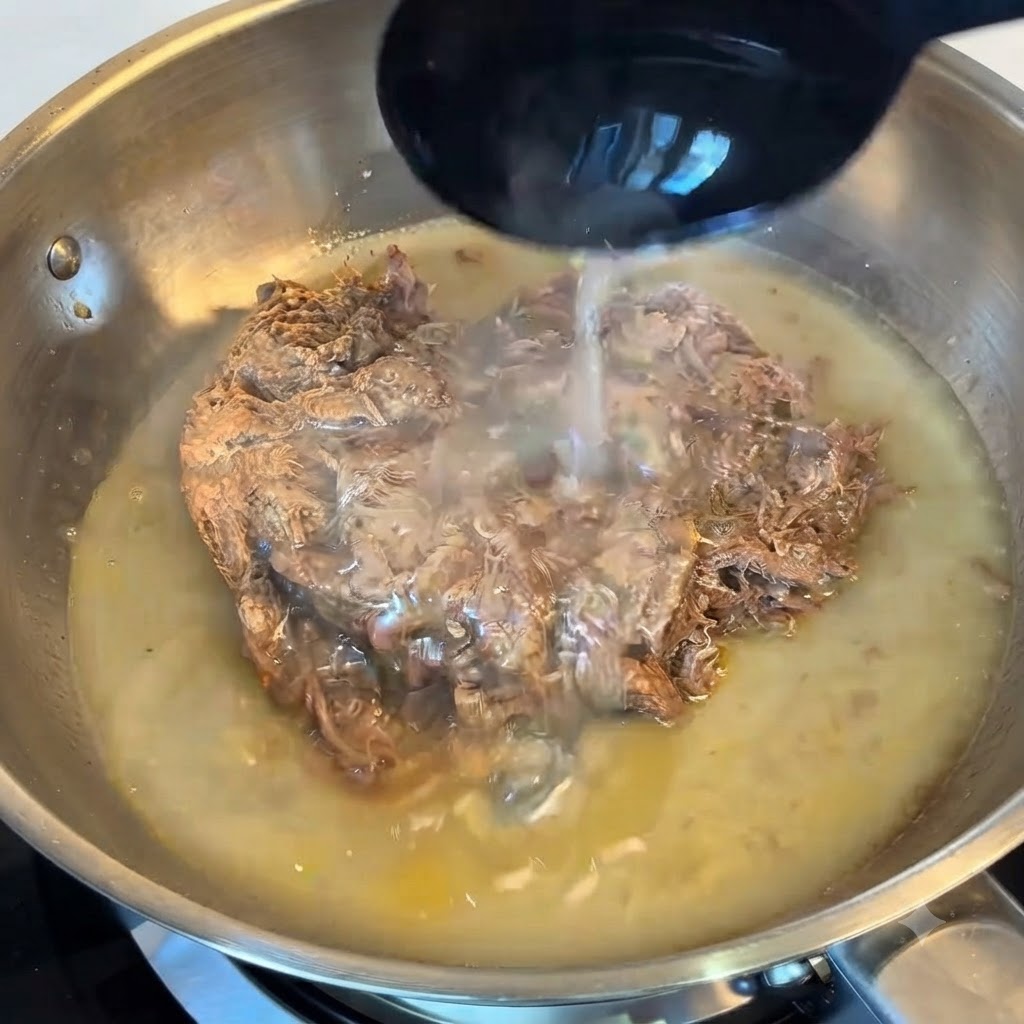 A ladle pouring starchy pasta water over the pulled beef in a stainless steel pan.