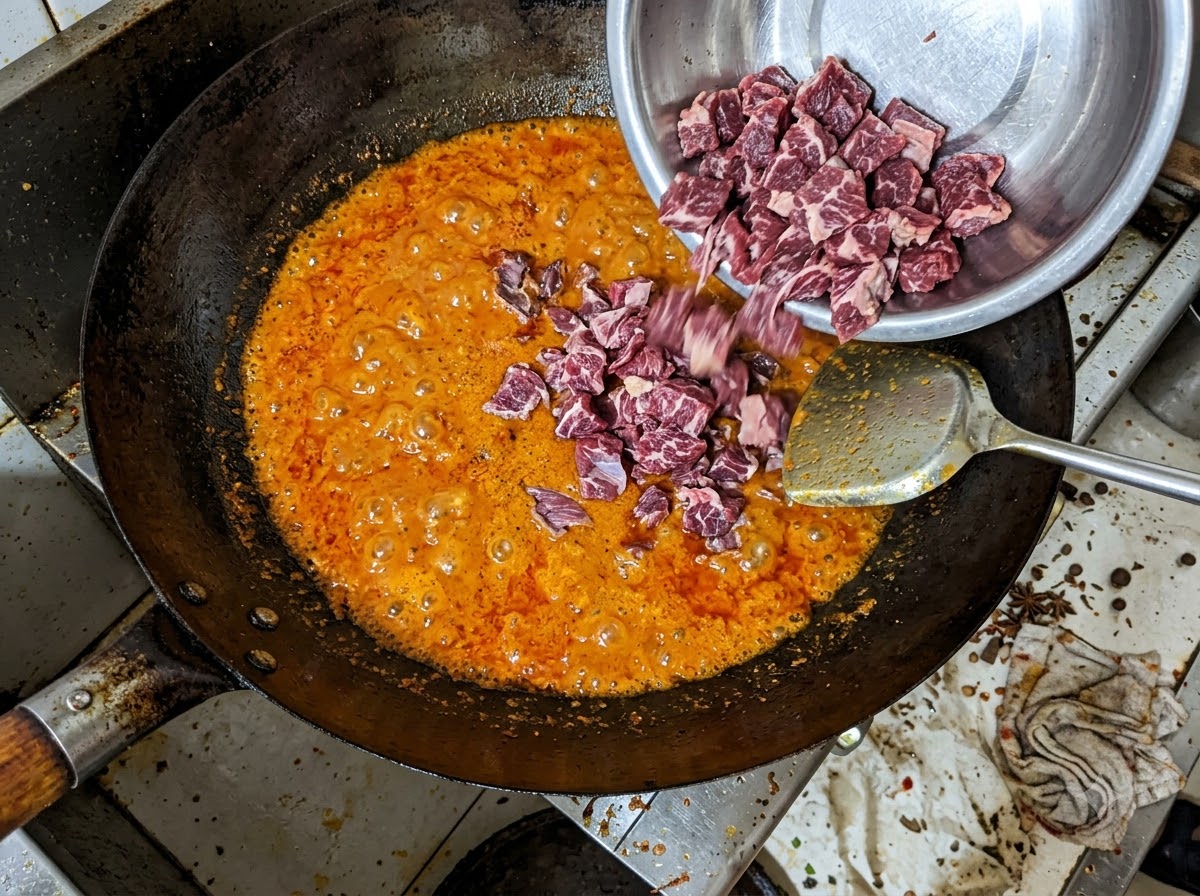 Chunks of beef brisket being poured from a metal bowl into a bubbling red chili oil mixture in a wok.
