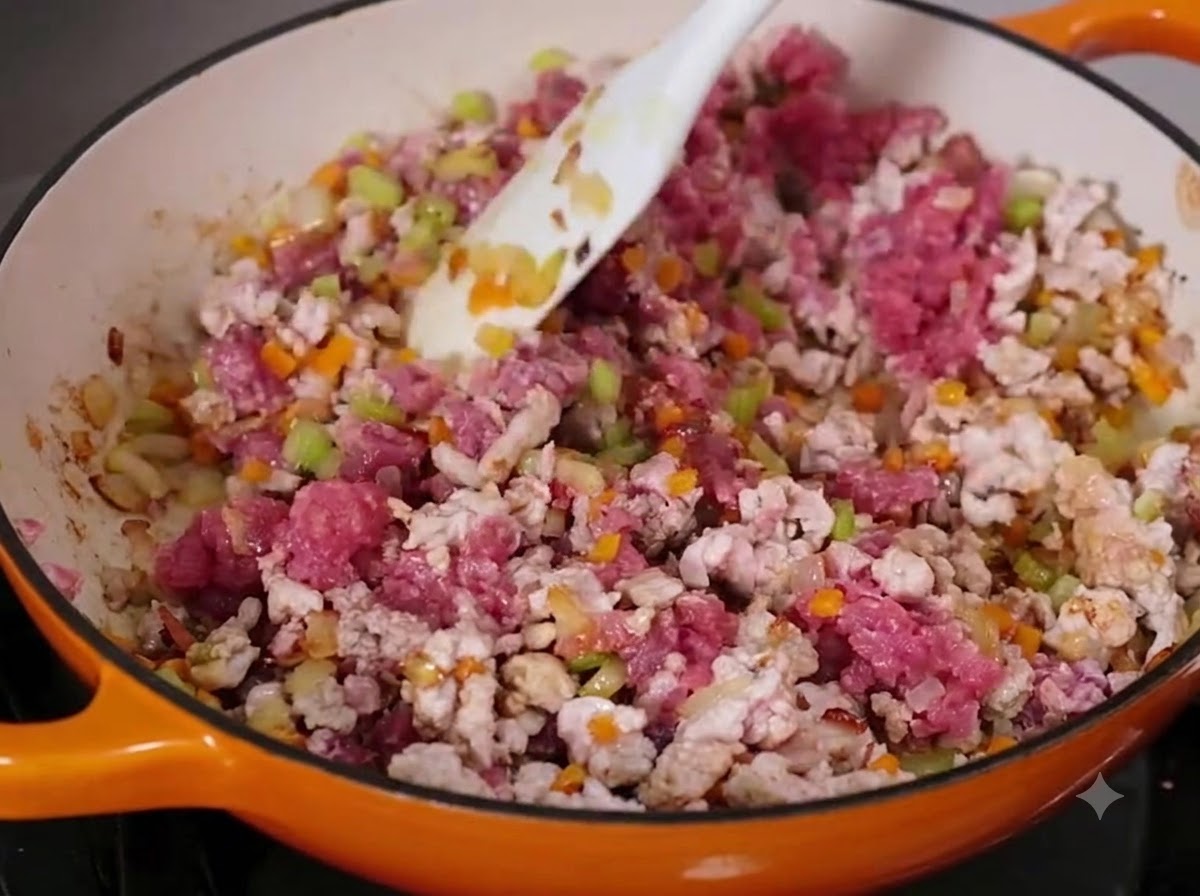 Ground beef and pork being mixed with diced vegetables in a large pan using a white spatula.