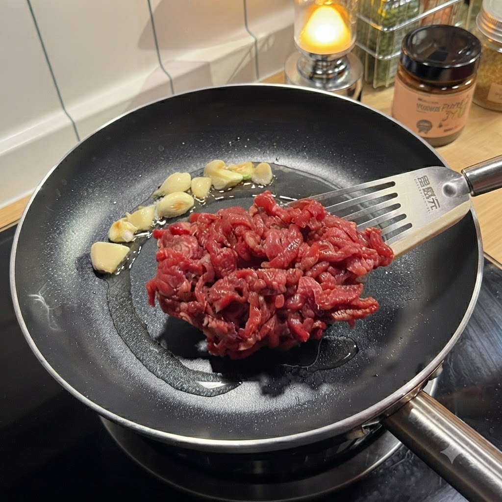 Seasoned raw beef strips being placed with a spatula into a hot pan containing sautéed garlic.
