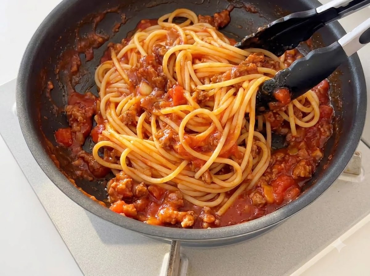 Cooked spaghetti noodles being tossed with thickened tomato meat sauce in a skillet using black silicone tongs.