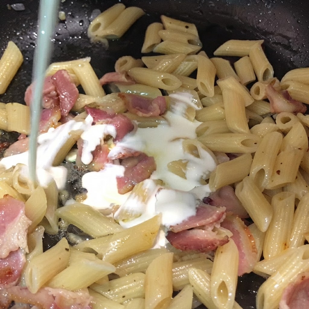 Milk and heavy cream being poured from above into a pan filled with penne pasta and bacon pieces.