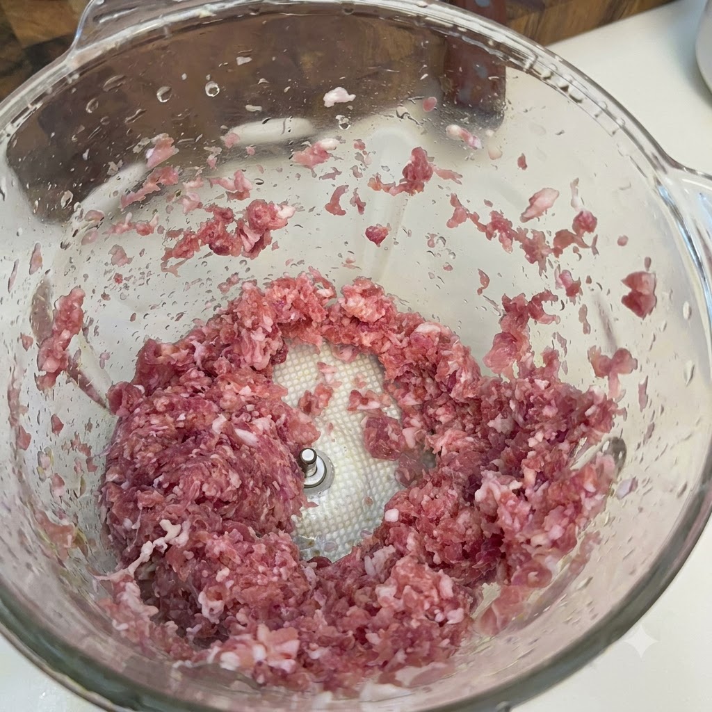 Minced pork or beef pieces loaded inside a glass food processor bowl.