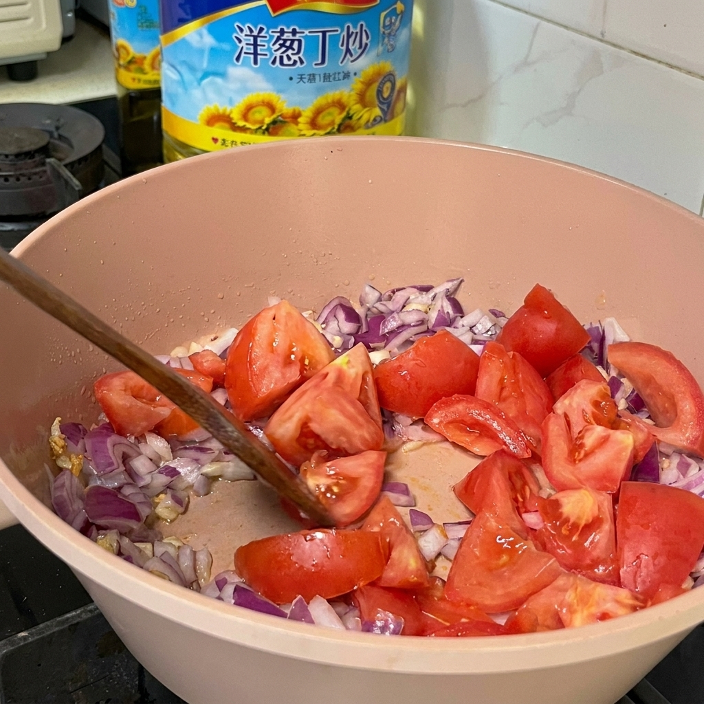 Fresh tomato chunks being mixed with diced red onions and garlic in a pink cooking pan using a wooden spatula.