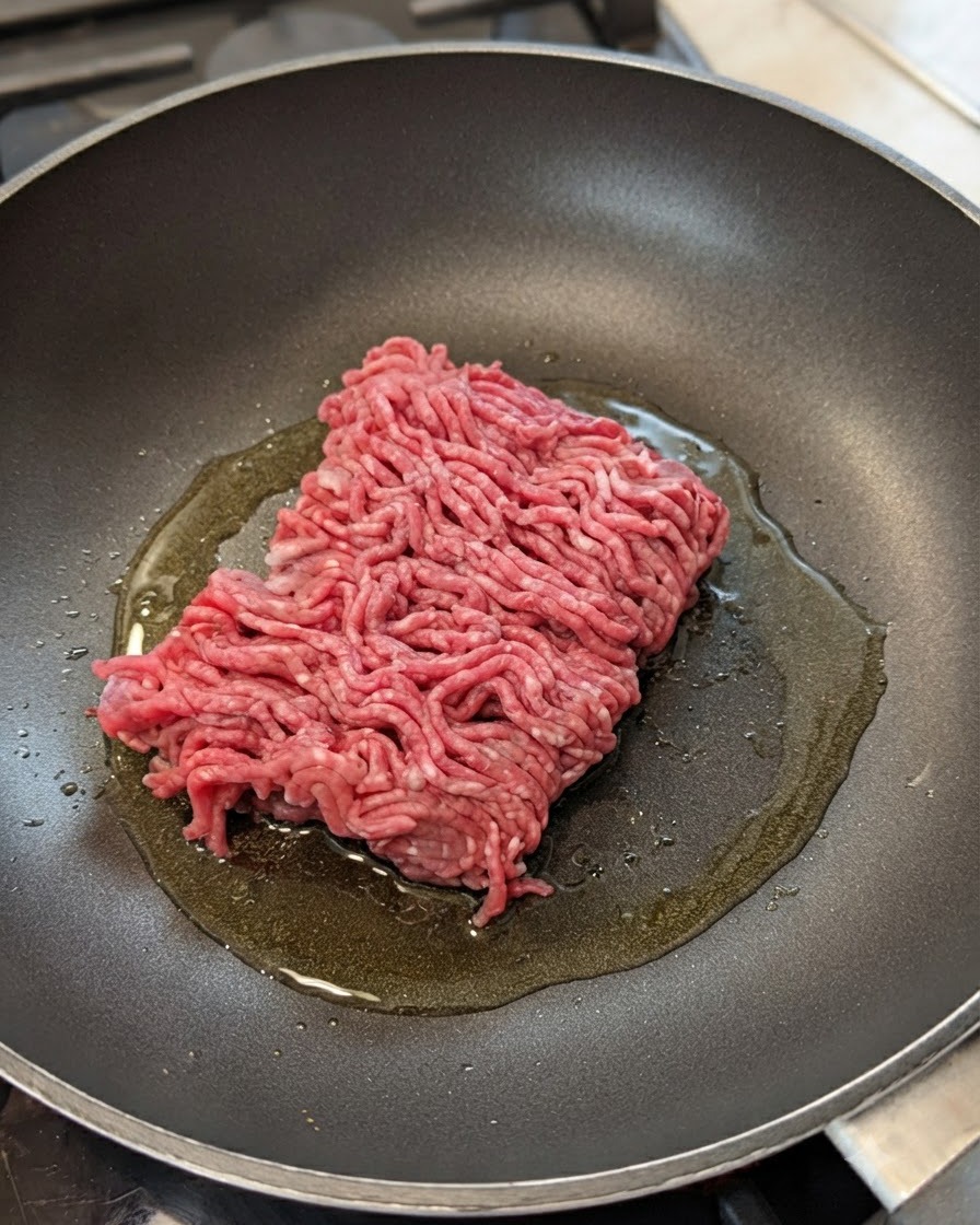 Raw ground beef sitting in a frying pan with oil, onions, and garlic.