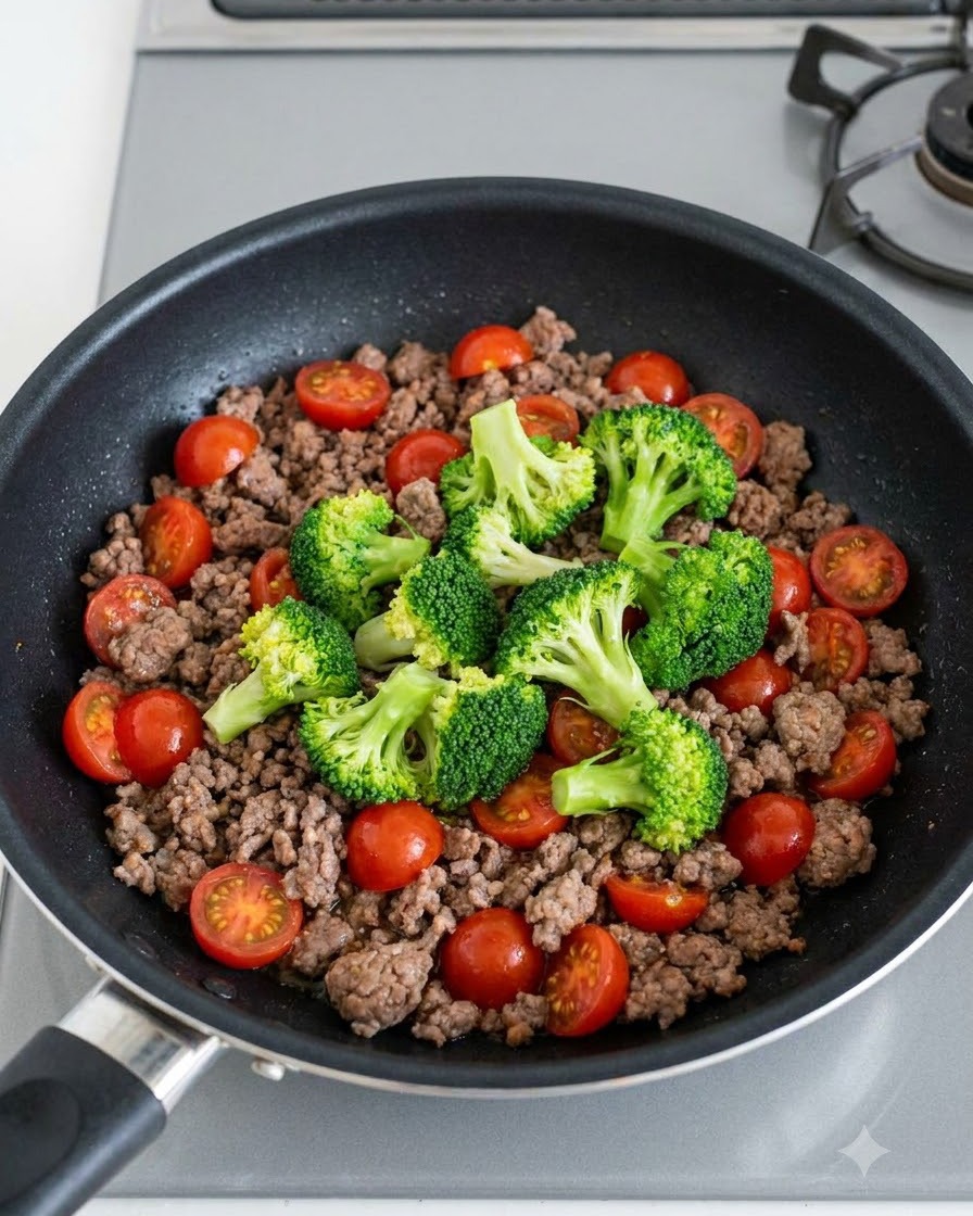 Green broccoli florets and bright red cherry tomatoes scattered over cooked ground beef in a pan.