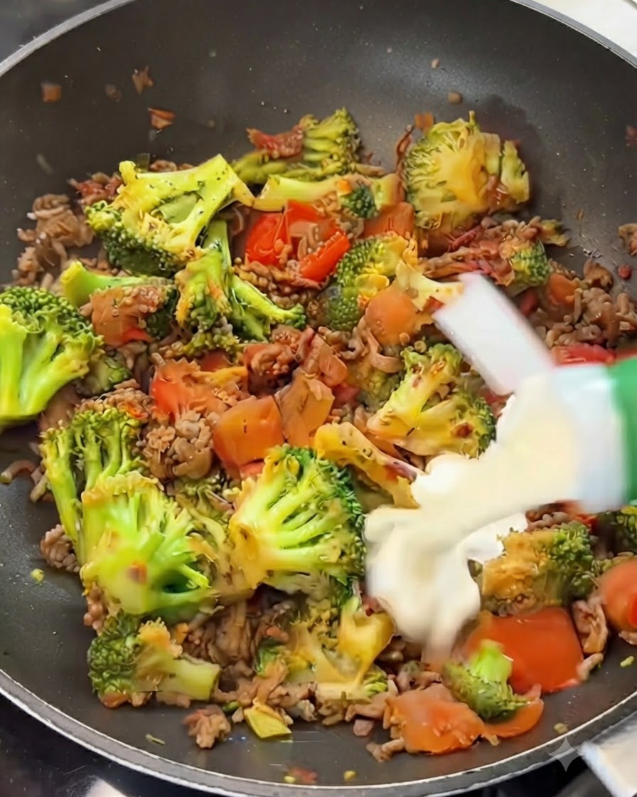 Heavy cream being poured from a carton onto a mixture of beef, broccoli, and tomatoes in a pan.