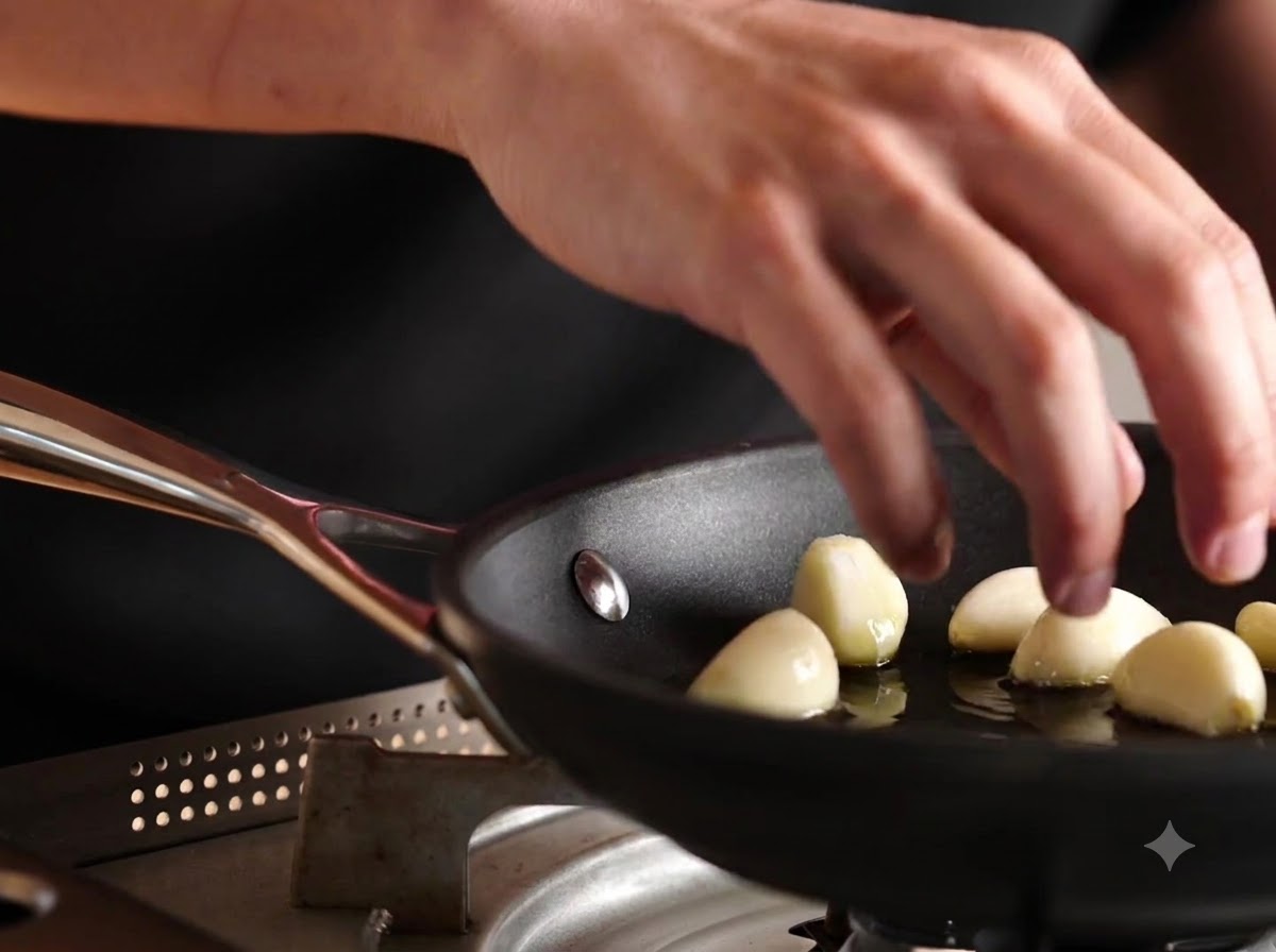 Whole peeled garlic cloves being dropped into a pan with olive oil.
