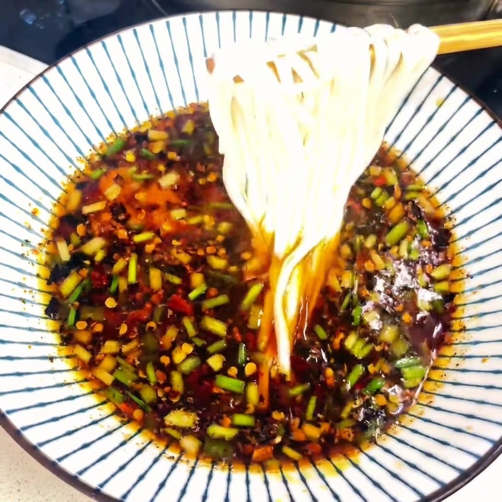 Chopsticks lifting a portion of cooked white noodles out of a bowl filled with seasoned broth.