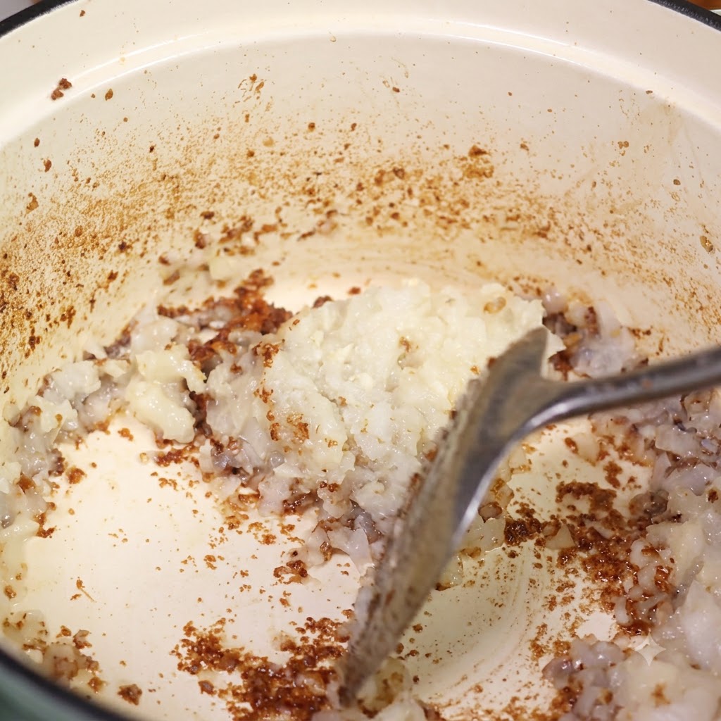 Minced onion being mixed into the pot over the browned fond using a spatula.