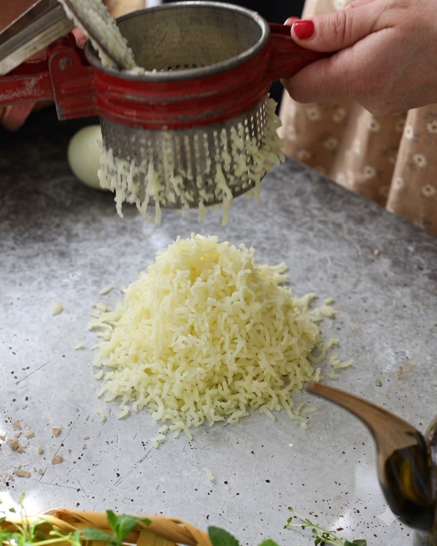 Hot boiled potatoes being pressed through a red potato ricer into a fluffy pile on the tabletop.