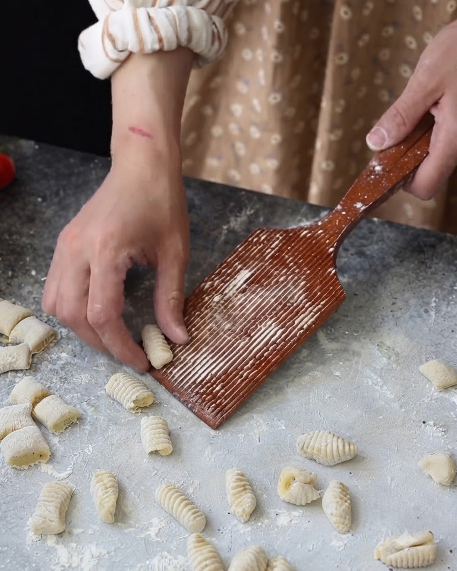 A hand rolling a small dough piece over a wooden grooved gnocchi board.