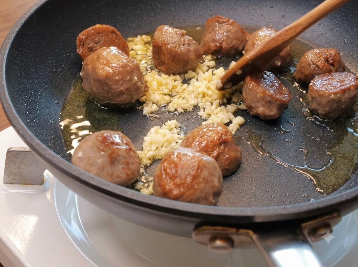 Minced garlic scattered among browning beef ball halves in a skillet with oil.