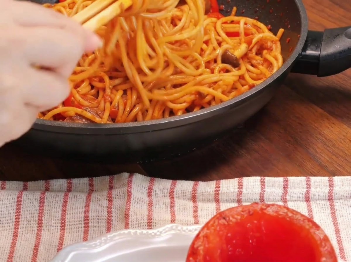 A close-up of a hand using chopsticks to twist spaghetti noodles inside a skillet next to a hollowed roasted tomato on a plate.