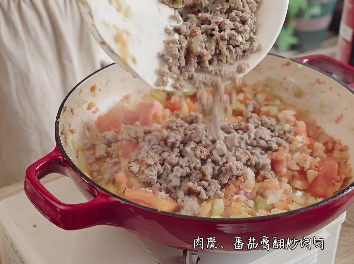 Ground meat being poured from a white plate into a red pot filled with chopped tomatoes, onions, celery, and carrots.