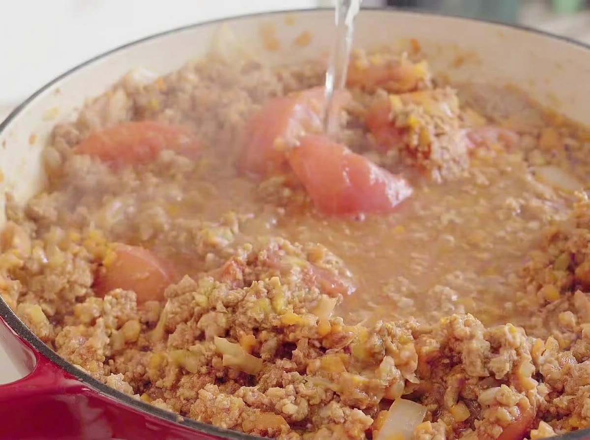 Water being poured from above into a large red pot containing the simmering meat, tomato, and vegetable mixture.