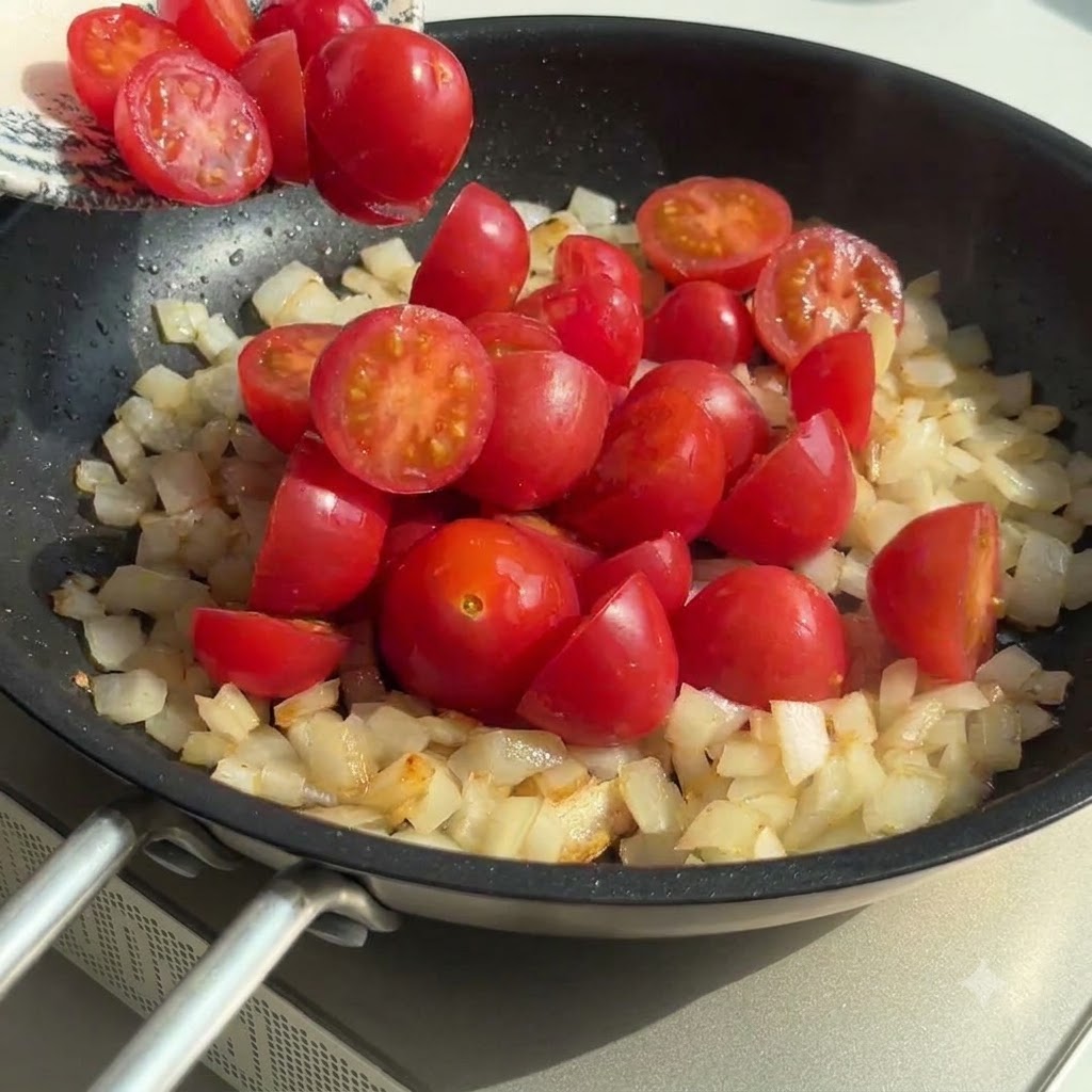 Halved cherry tomatoes being added to the pan with sautéed onions and garlic, stirred together.