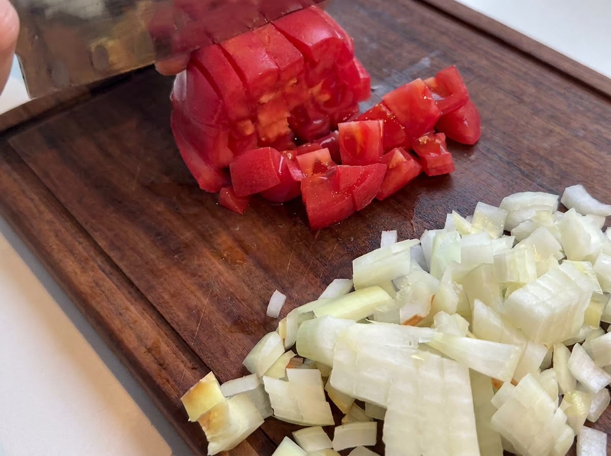 Fresh red tomato being diced with a kitchen knife on a wooden cutting board next to chopped onions.