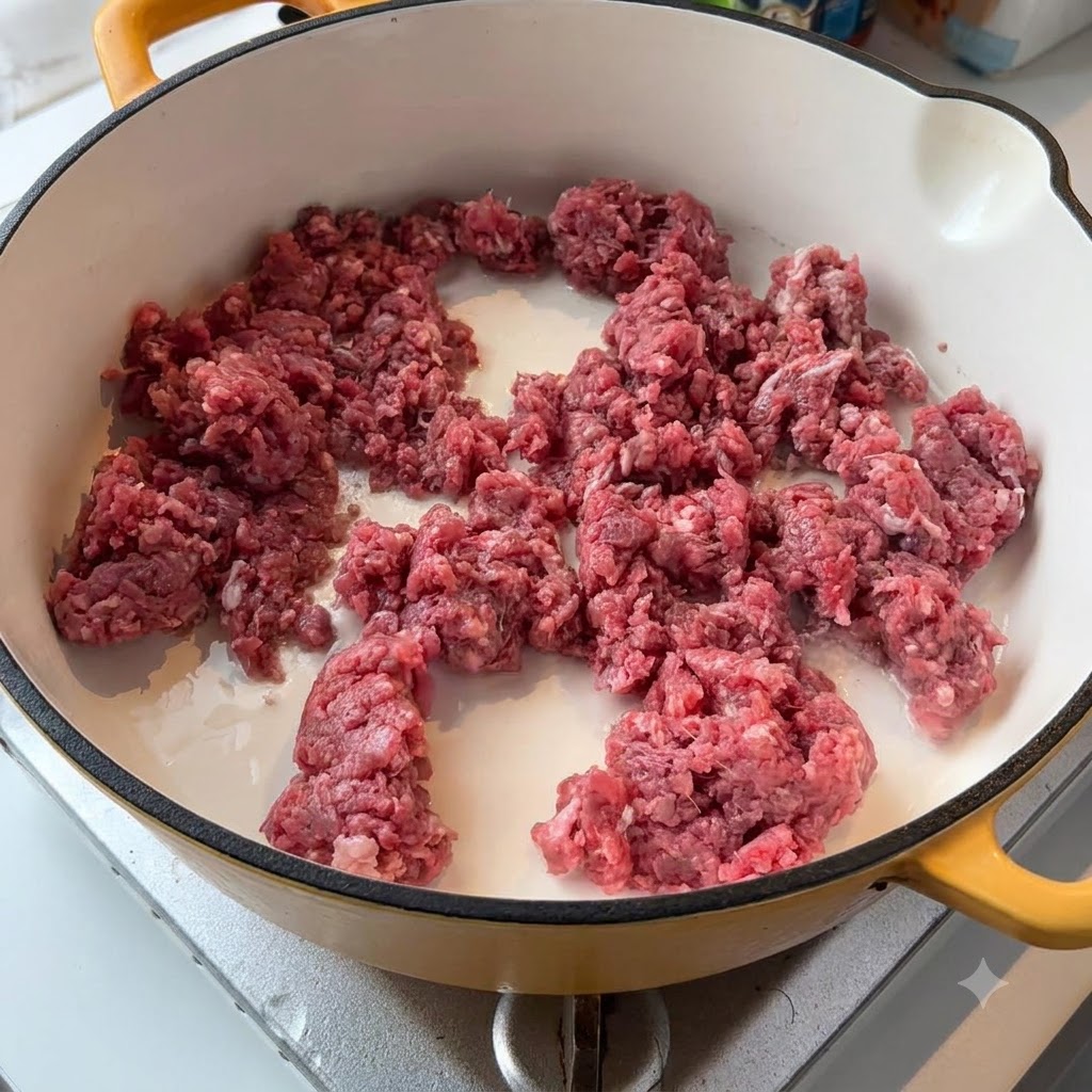 Raw minced beef and white onions sizzling in a light-colored pan on the stove.