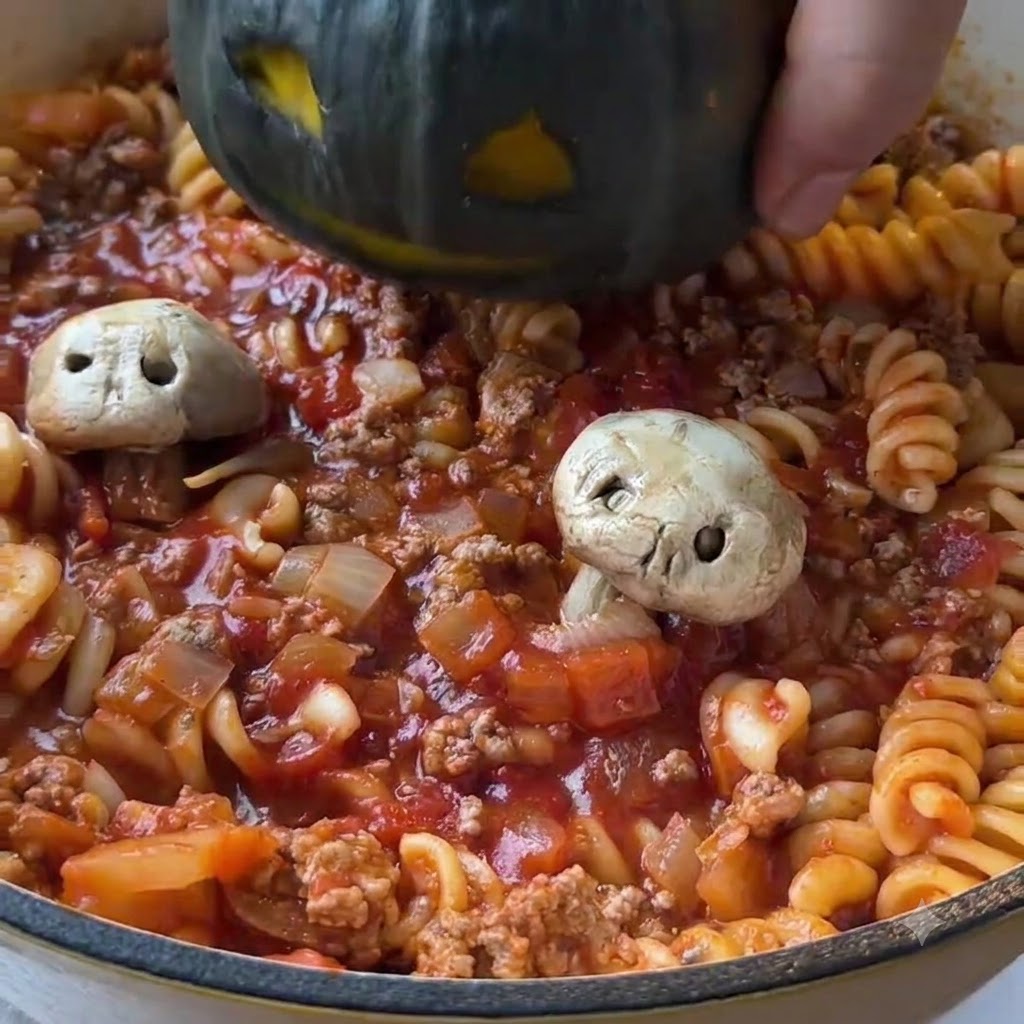A carved dark green mini pumpkin being lowered into the center of a pan filled with cooked tomato pasta and minced meat sauce.