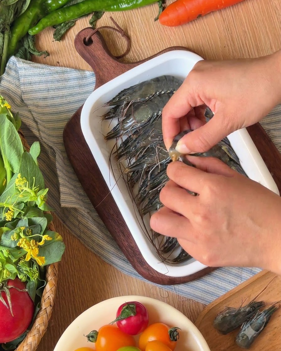 Hands peeling the shells and removing the heads from raw black tiger prawns in a white ceramic tray.