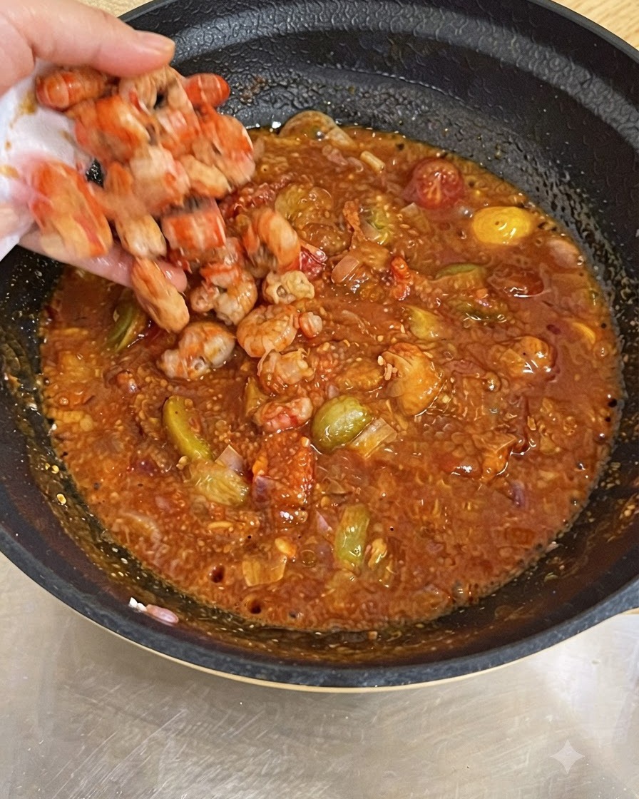 A hand dropping cooked shrimp into a pan filled with bubbling red tomato sauce.
