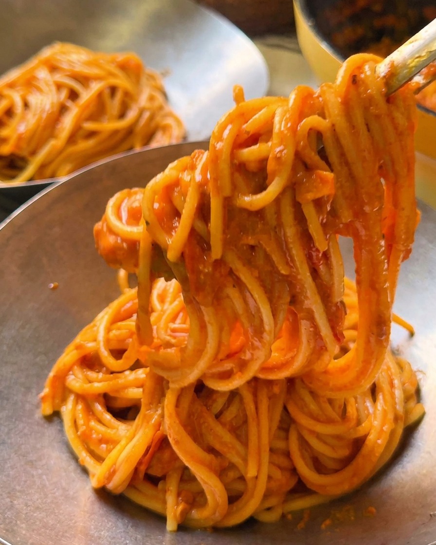 Tongs lifting a portion of creamy tomato spaghetti into a polished metal serving bowl.