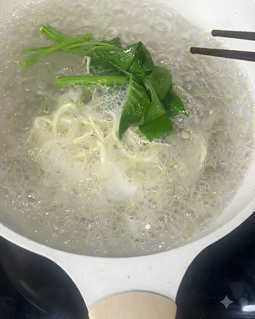 Chopsticks stirring thin white noodles and green leafy vegetables boiling in water.