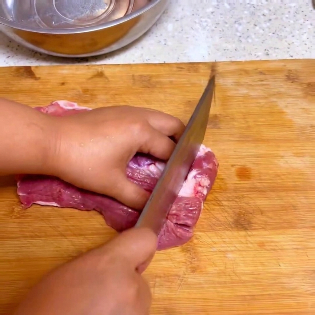 A kitchen knife slicing fresh raw beef into large chunks on a wooden cutting board.