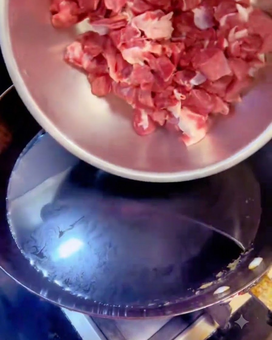 Beef cubes being poured from a steel bowl into a pot of boiling water with aromatics.