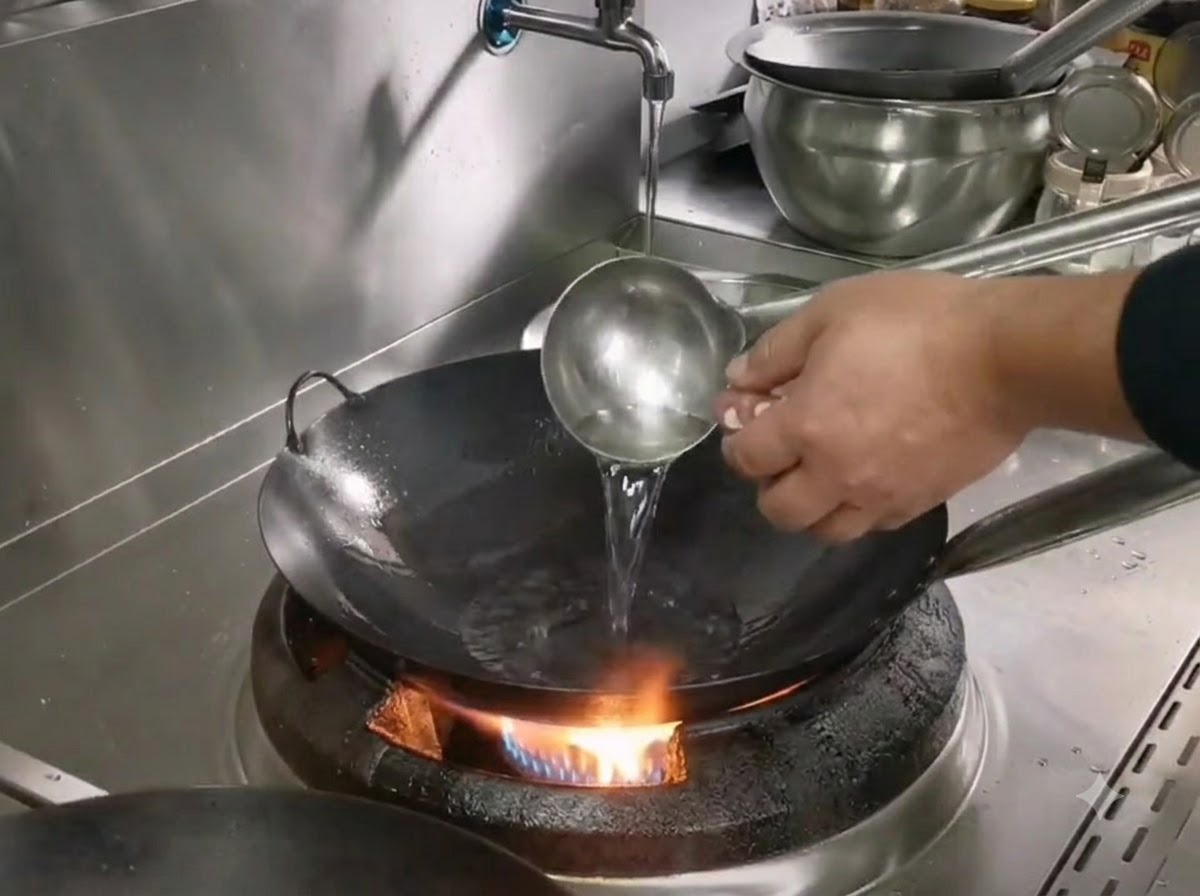 A ladle pouring oil into a hot iron wok over a gas burner while preparing to cook shredded pork.