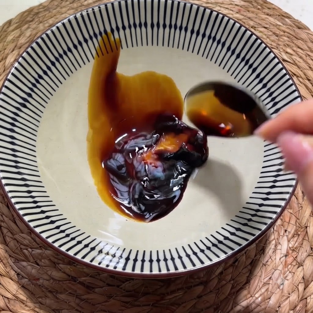 A hand pouring dark soy sauce from a spoon into a patterned ceramic bowl containing a dollop of oyster sauce.