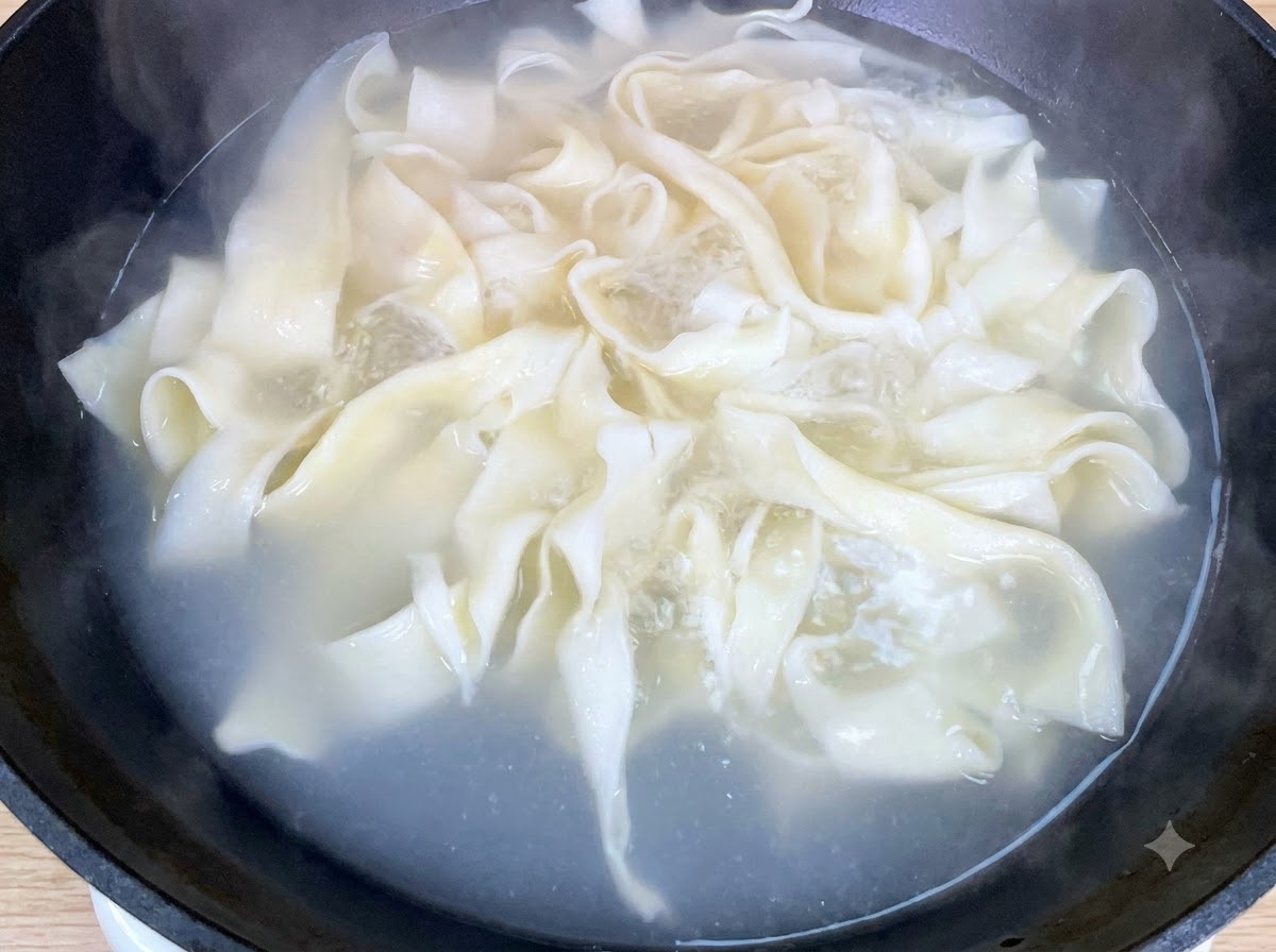 Fresh baby bok choy boiling alongside wide noodles in a pan.