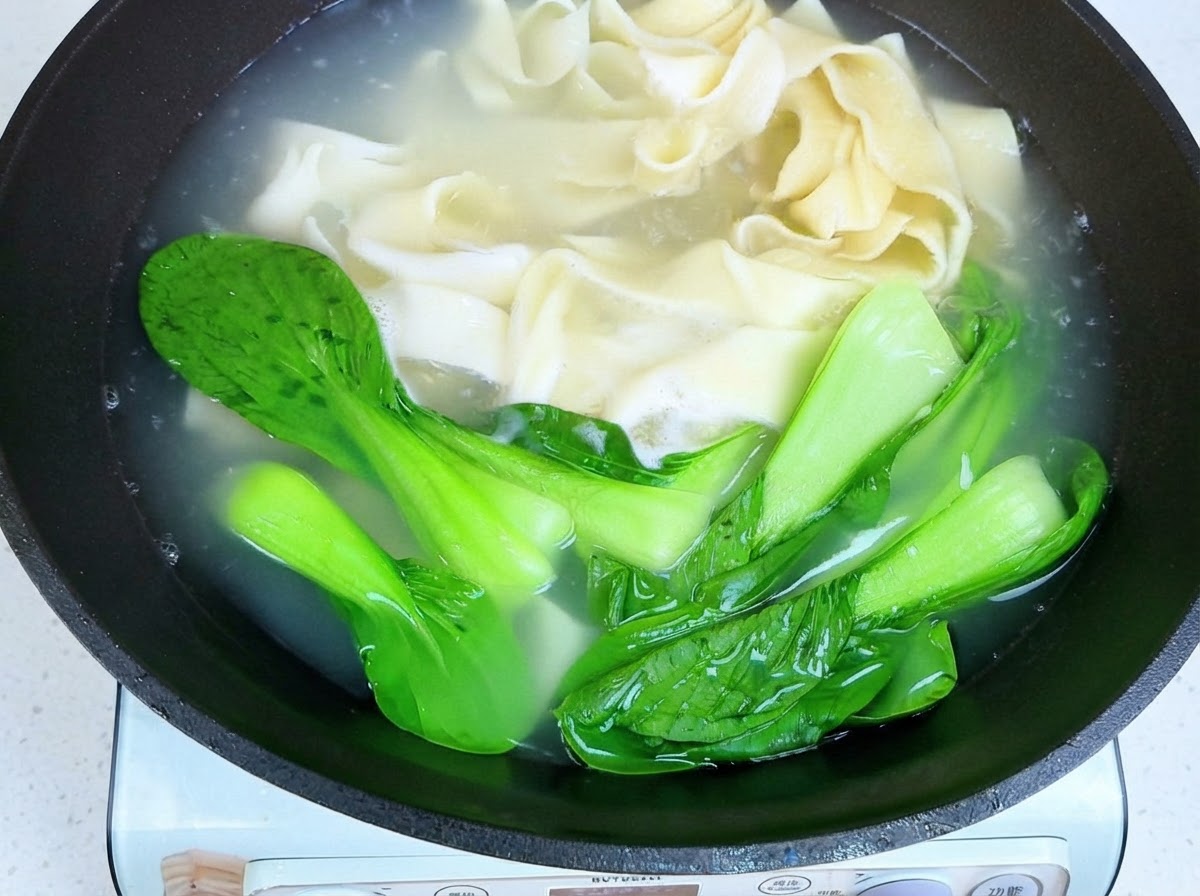 Chopsticks lifting cooked wide noodles and bok choy out of hot water.