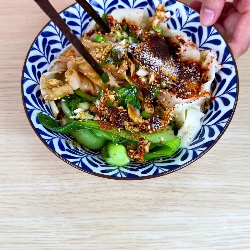 Chopsticks mixing the wide wheat noodles, bok choy, and chili oil sauce together inside the ceramic bowl.