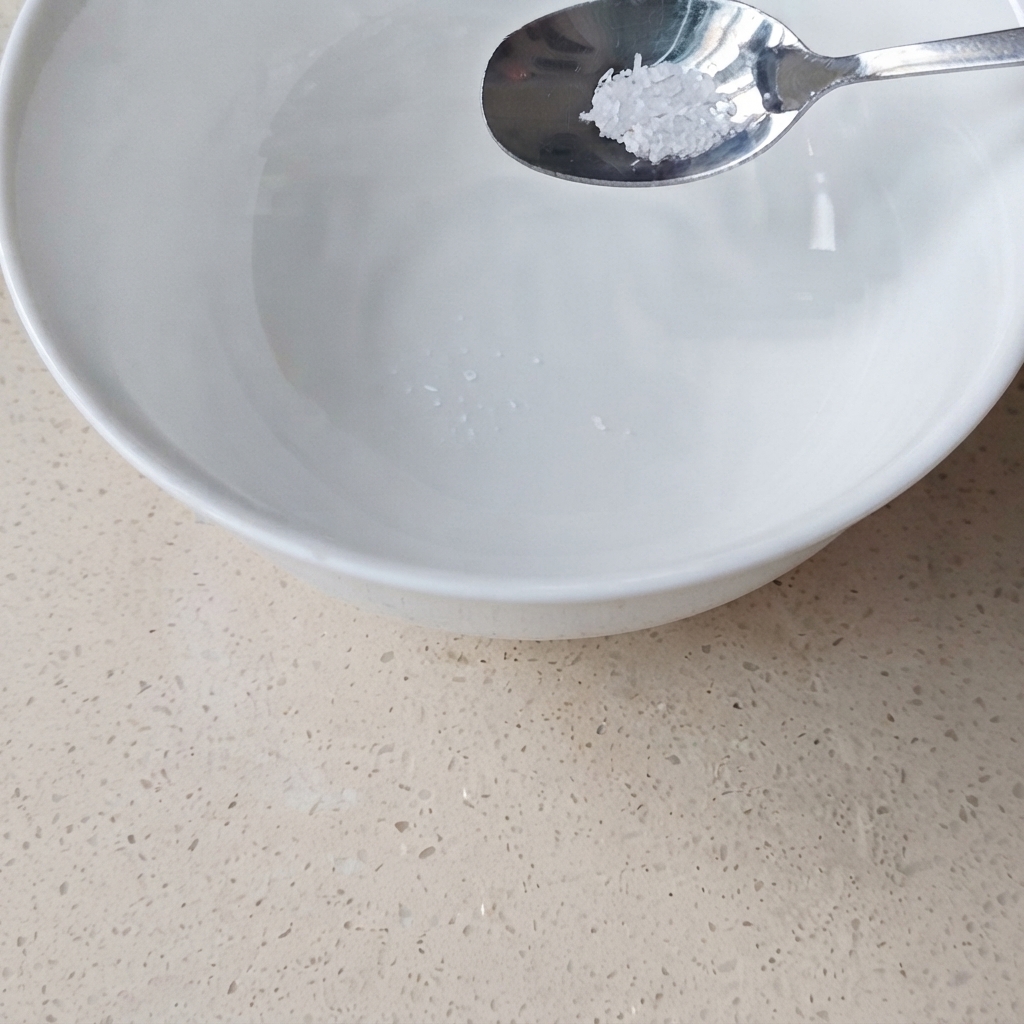 A metal spoon holding a small amount of MSG positioned over a white ceramic bowl containing water.