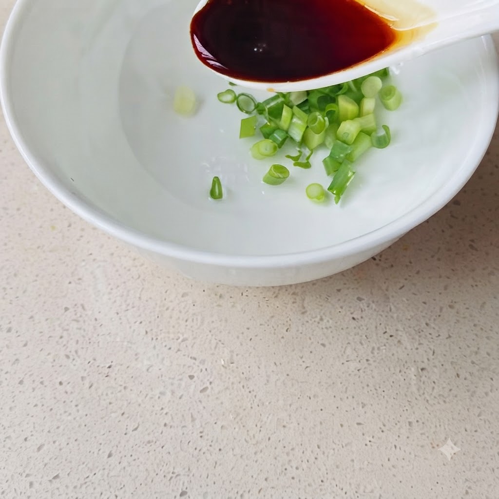 A white spoon pouring dark soy sauce into a white bowl containing chopped spring onions and water.