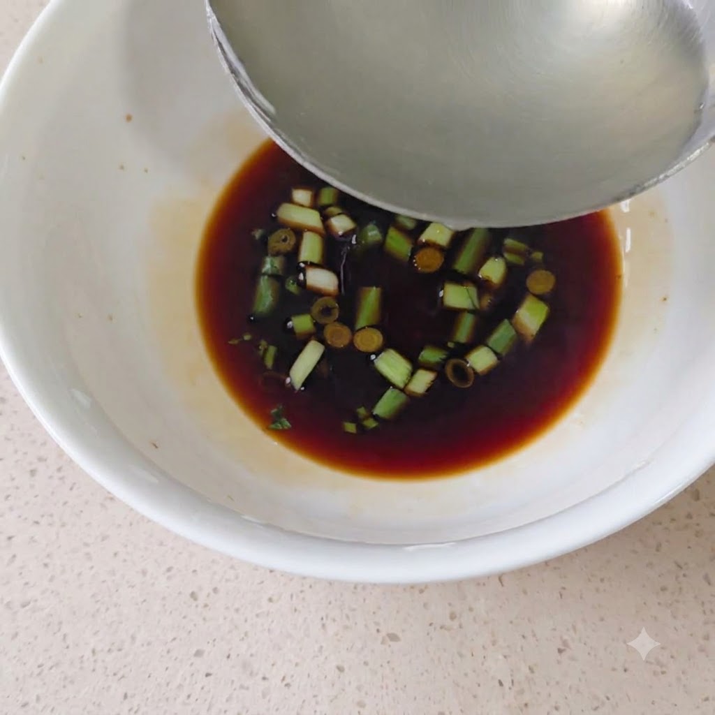 A metal ladle pouring hot broth into a white bowl filled with soy sauce and chopped spring onions.