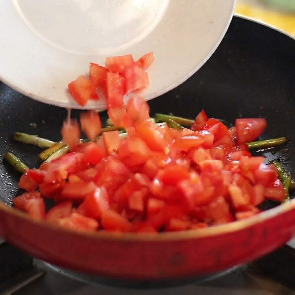 Diced red tomatoes sliding from a white plate into a hot pan with sautéed scallions.