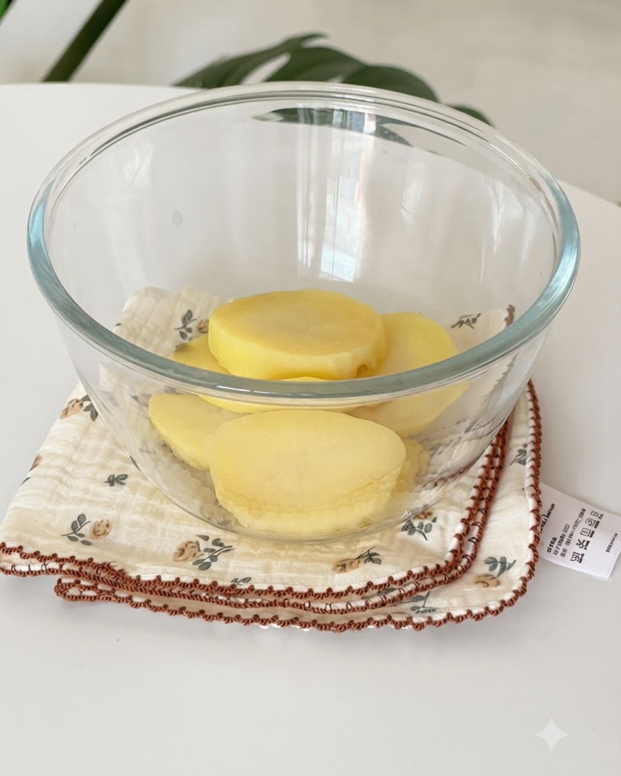 Slices of steamed yellow potatoes resting inside a clear glass bowl on top of a cloth.