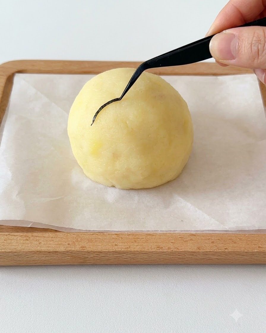 A hand holding black tweezers placing a thin curved piece of nori seaweed onto a spherical potato head.