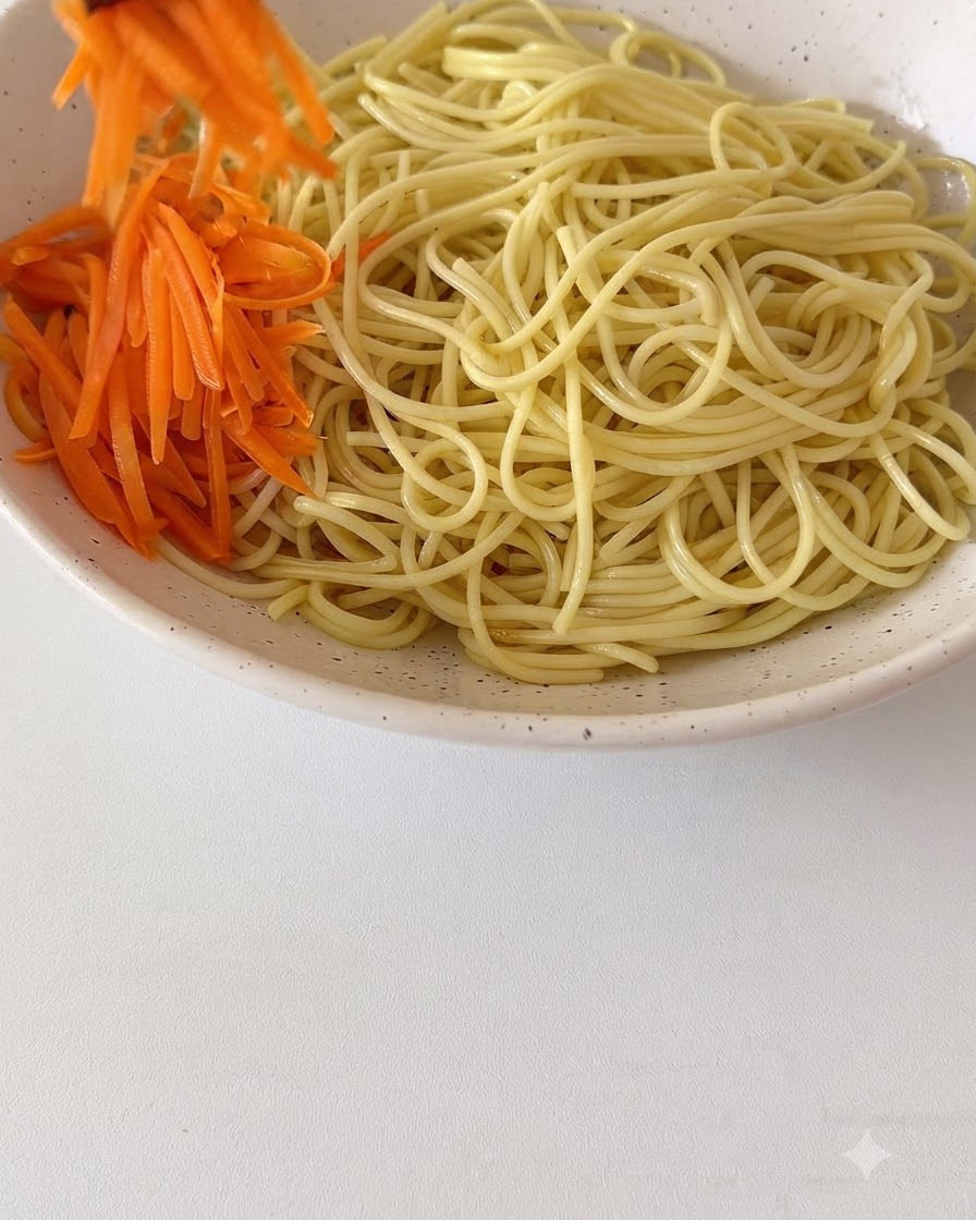 Fresh shredded carrots and other toppings being arranged on top of the cooked noodles in a large ceramic bowl.