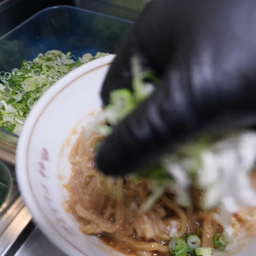 A gloved hand scattering chopped green onions and chives over the seasoned noodles in the bowl.