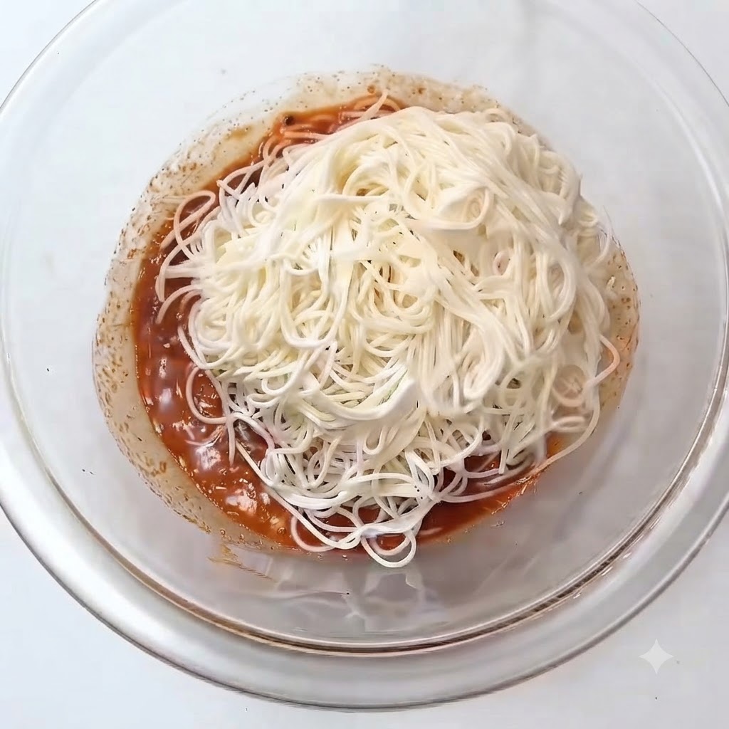 A pile of white cooked somen noodles placed on top of the reddish sauce inside a glass mixing bowl.