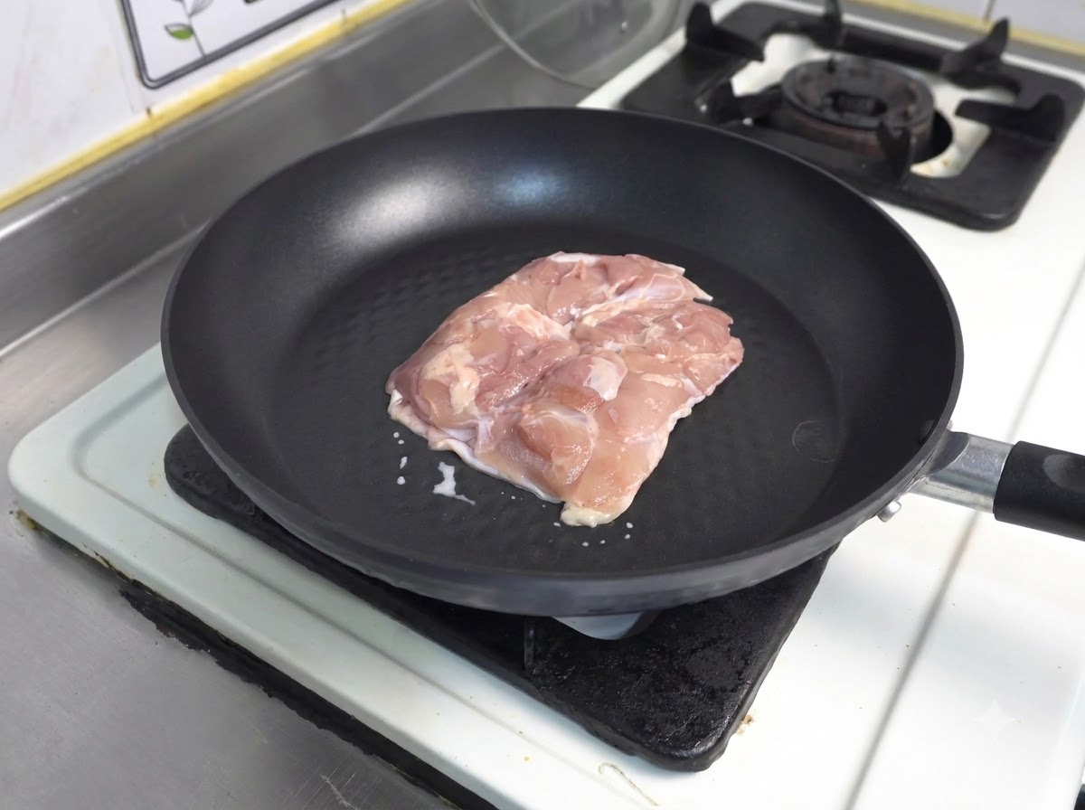 A piece of raw chicken thigh skin-side down in a dark non-stick frying pan.